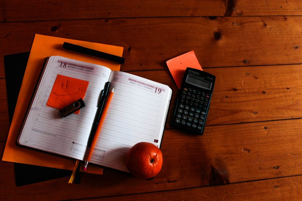 End-of-summer tax checkup reminder with an open planner with a smiley face sticky note, pen, pencil, a USB drive, an orange, calculator, and papers on a wooden surface.