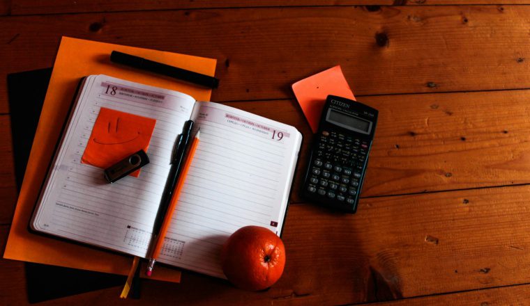 An open planner with a smiley face sticky note, pen, pencil, a USB drive, an orange, calculator, and papers on a wooden surface.